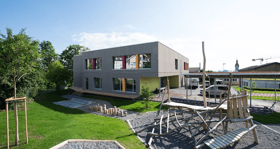 Zweigeschossiger Holzbau mit Spielplatz, Bäumen und gedeckten Parkplätzen